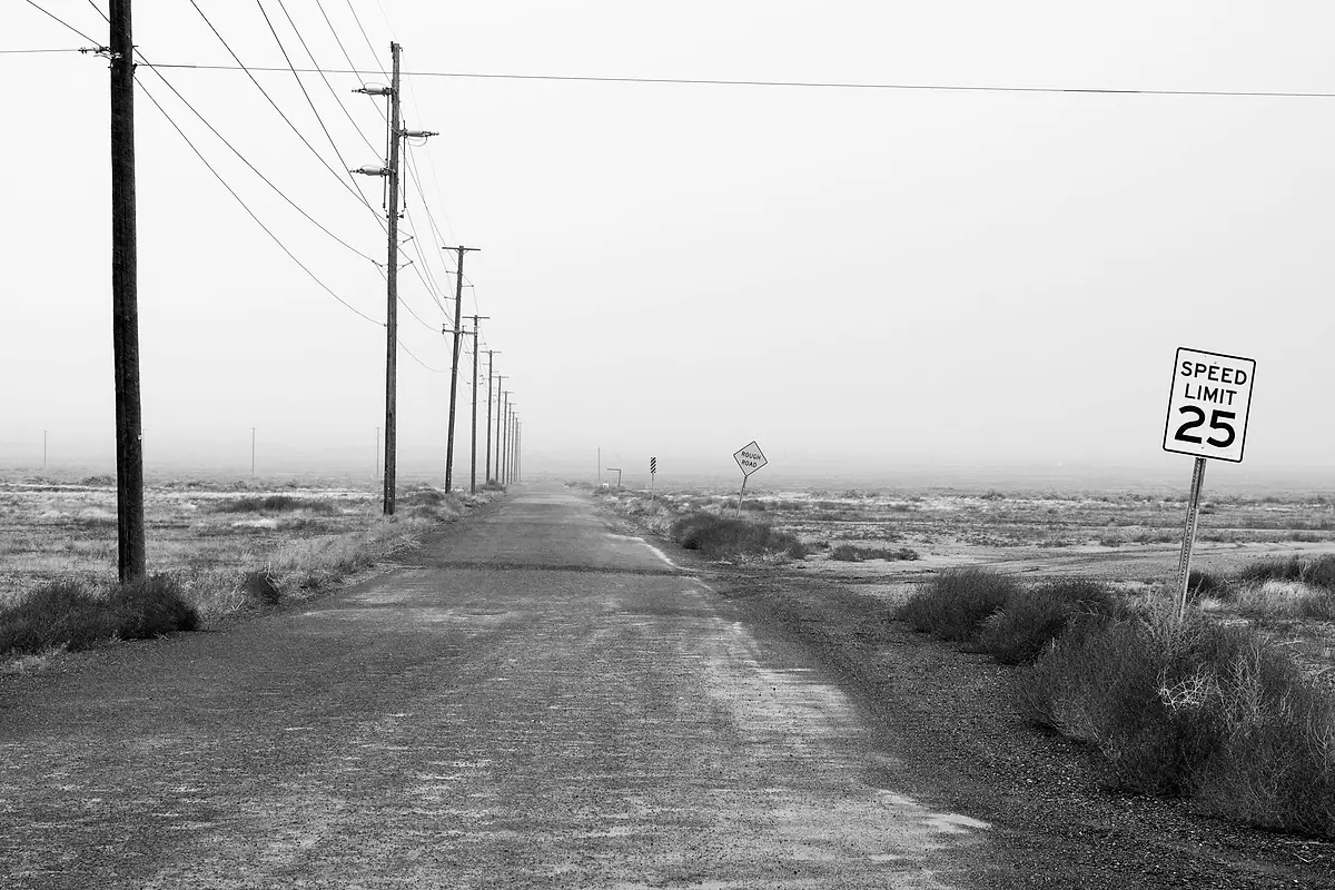 Black-and-white photo of an empty rural road lined with utility poles and a 25 mph speed limit sign.